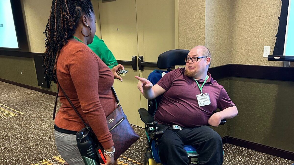 Nicolas Comstock smiling, wearing a Nick Speaks branded shirt, in his power wheelchair.